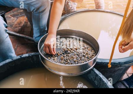 Child hand picking pebbles at the sieve at archaeological excavations or extraction of gold and other gems at the prospecting site. Muddy water in bac Stock Photo