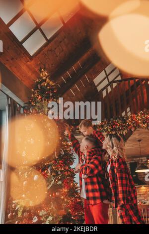 Smile active parents with small son in red checkered sleepwears waiting Santa indoor. Stylish family love laughing hugs kisses at home. Celebrate New Stock Photo