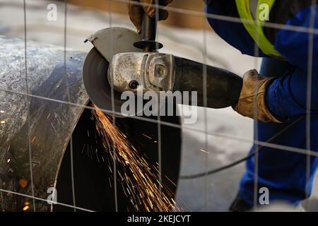 Shallow depth of field (selective focus) details with a worker using an angle grinder on an industrial metallic pipeline. Stock Photo
