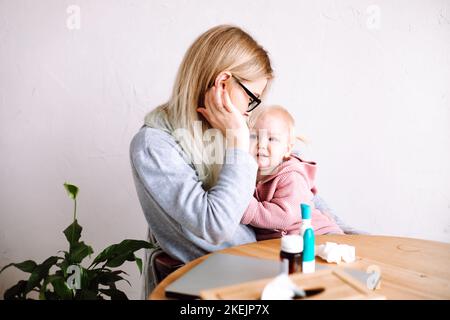 Side view of young mother woman sitting at table, holding little sad baby girl, calming down child at home. Sick time. Stock Photo