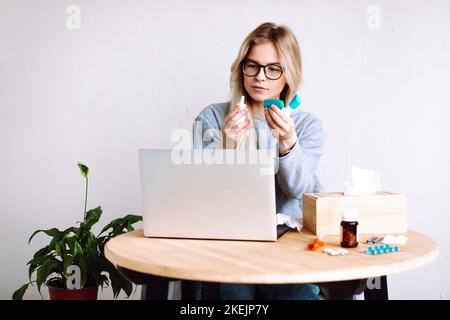 Portrait of young woman sitting near laptop box with paper napkins, holding bottles for sore throat, nasal spray bottle. Stock Photo