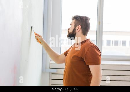Young man using spatula and plastering of wall with white fresh finishing putty. Closeup. Repair work of home. Side view. Stock Photo