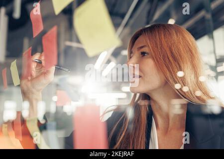 Businesswoman writing on sticky notes on glass wall while working in office Stock Photo