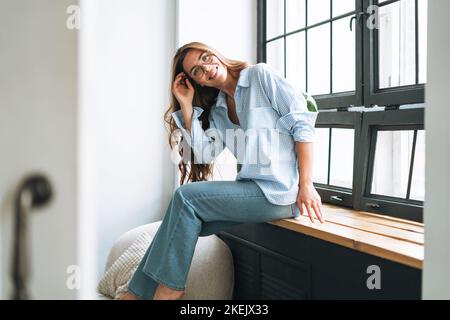 Portrait of young smiling woman with long hair sitting on window sill  in modern office Stock Photo