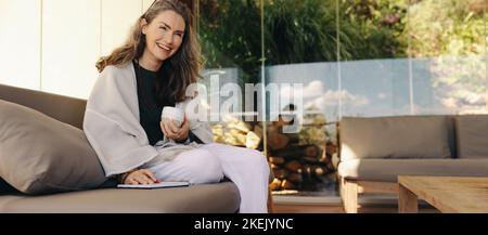 Happy senior woman journaling with a cup of tea at home. Mature woman smiling cheerfully while sitting on a couch. Senior woman enjoying a serene morn Stock Photo