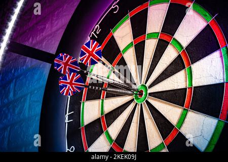 Three darts with uk flag flights in triple double bullseye on professional sisal steeldart board in colorful LED illumination on white stone wall. per Stock Photo
