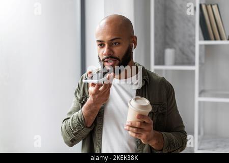 Latin man recording voice message on smartphone and drinking coffee from takeaway cup, enjoying modern technologies Stock Photo