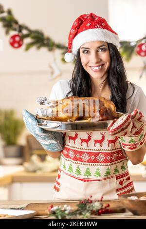 Satisfied young woman in a Christmas apron and Santa hat holding a roasting pan with a roast goose or turkey. Stock Photo