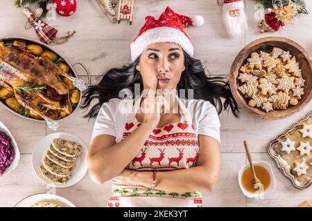 Pensive female cook in a Christmas apron and Santa hat,lying on the floor, surrounded by gingerbread, Linz cakes, on the other side: roast goose or tu Stock Photo