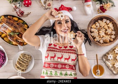 A cheerful cook in a Christmas apron is lying on the ground holding a Linzer pastry and a gingerbread star surrounded by traditional holiday dishes an Stock Photo