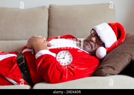 Older man dressed as Santa Claus sleeping on the sofa next to the alarm clock. Christmas time. Stock Photo