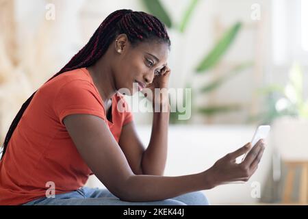 Worried Black Woman Looking At Smartphone Screen At Home, Side View Stock Photo