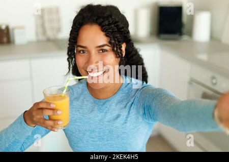 Positive african american fit woman drinking fresh juice from straw and taking selfie, standing in kitchen interior Stock Photo