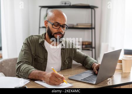 Focused mature latin man taking notes while using laptop, working or learning distantly online, sitting at desk at home Stock Photo