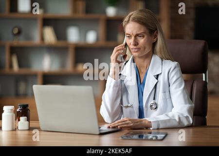 Worried Doctor Woman Using Laptop And Talking On Cellphone At Workplace Stock Photo
