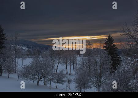 The orange rays of the sun break through the dark gray clouds and illuminate the snow-covered mountains and forests. Stock Photo