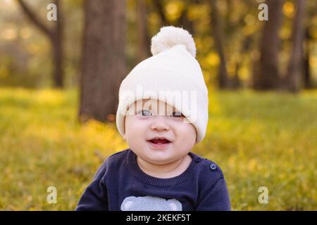 baby in white knitted hat sits on grass in Park against background of autumn trees Stock Photo