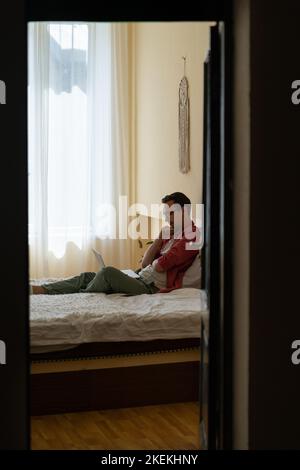 Pensive young man student sits on bed with laptop on knees watching education video from college  Stock Photo
