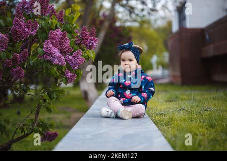 A little girl stands near a lush bush of lilacs, she smiles and sniffs purple flowers Stock Photo