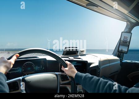 Trucker drives a modern truck across the bridge. View from the cabin to the sea and wind turbines. Stock Photo