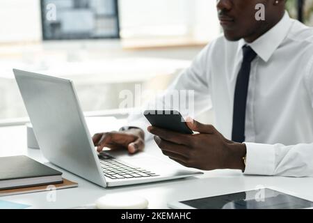 Young African American using smartphone and laptop at his work while sitting at table at office Stock Photo