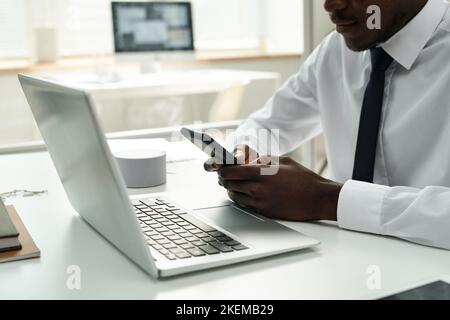 African American businessman typing a message on his smartphone while working on laptop at his workplace at office Stock Photo