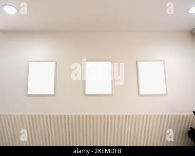 Three empty white blank pictures of brown wood photo frames set hanging on beige wall background in coffee shop. Mockup vintage frames on minimal clea Stock Photo