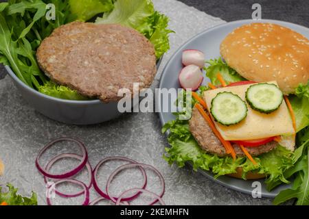 Beef burger with greens in bowl. Cheeseburger with cucumber, greens and carrots. Chopped onion on table.  Black background. Stock Photo