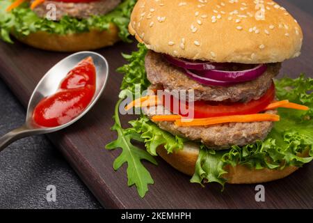 Beef burger with greens, chopped onions and carrots on board. Tomato sauce in bowl. Close up. Black background. Stock Photo