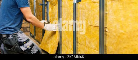 house wall insulation. construction worker installing glass wool into the metal profile frame indoors. banner with copy space Stock Photo