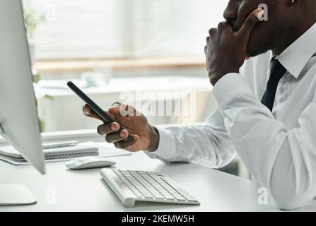 Close-up of African American businessman using his smartphone while sitting at table in front of monitor at office Stock Photo