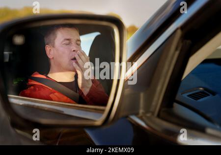 A tired driver yawns while covering his mouth with his hand while sitting at the wheel. View of the driver through the side view mirror. Fuzzy image. Stock Photo