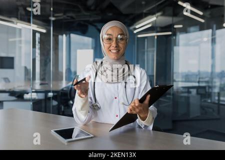 Online doctor's appointment. A young beautiful woman doctor, a Muslim woman, consults a patient remotely through a video call. He looks into the camera, explains, smiles, writes down complaints. Stock Photo
