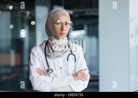 Portrait of a young Muslim woman doctor. She is standing in the hall of the hospital dressed in a hijab, wearing glasses and a stethoscope, looking seriously into the camera, her arms crossed. Stock Photo