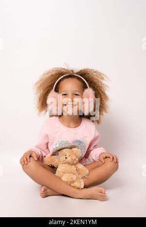 Little child girl wearing earmuffs is hugging teddy bear isolated over white background. Stock Photo