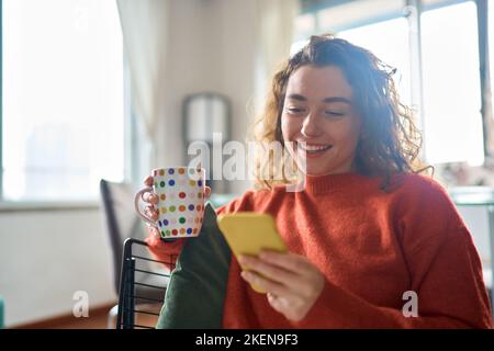 Young happy woman using mobile phone drinking coffee relaxing at home. Stock Photo