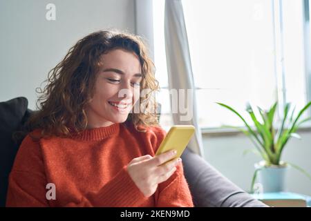 Smiling young woman sitting on couch using cell phone doing online shopping. Stock Photo
