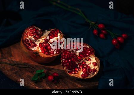 Pomegranate on a cutting board and rosehip branches on black table. Still life. Closeup. Stock Photo