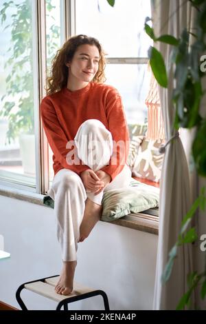 Happy serene young woman sitting relaxing at home looking through window. Stock Photo