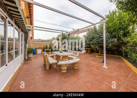 single-family house with a porch with a tiled patio and a sliding fabric gazebo over a stone table and benches Stock Photo