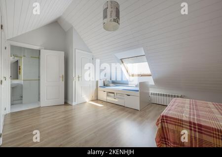 Bedroom with white cabinet doors, bed with striped bedspread, sloping ceilings and youthful bed with drawers, skylight in the ceiling and en-suite toi Stock Photo
