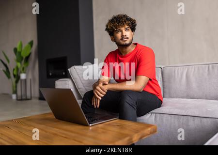 Side view of positive indian guy in casual outfit sitting on couch at home, using modern laptop, typing on computer keyboard and smiling, freelancer e Stock Photo