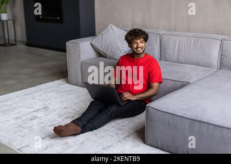 Young handsome indian man working on laptop sittin on floor in living room Stock Photo