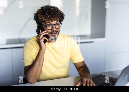Smiling young arab man freelancer talking on mobile phone and taking notes, working from home during isolation, copy space. Happy guy having conversat Stock Photo