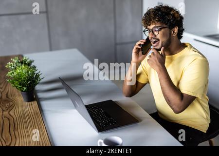 Smiling young arab man freelancer talking on mobile phone and taking notes, working from home during isolation, copy space. Happy guy having conversat Stock Photo
