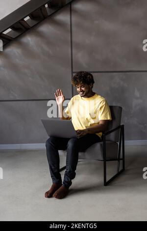 Young man freelancer is at home, sits in a chair and communicates online with a woman. A young man on a laptop uses a video call and video chat. Stock Photo