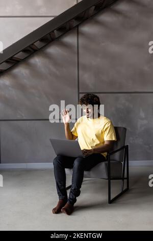 Young man freelancer is at home, sits in a chair and communicates online with a woman. A young man on a laptop uses a video call and video chat. Stock Photo