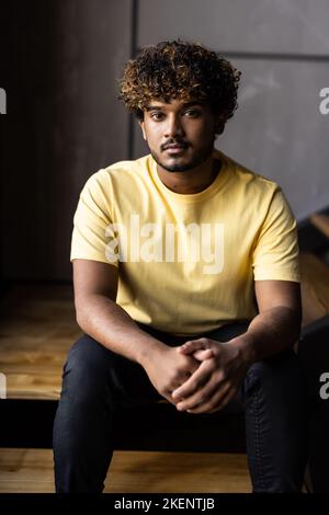 Thoughtful indian man user sitting at home on stairs relaxing wearing earbuds using smartphone thinking looking through window drinking coffee listeni Stock Photo