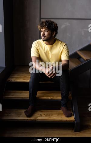 Thoughtful indian man user sitting at home on stairs relaxing wearing earbuds using smartphone thinking looking through window drinking coffee listeni Stock Photo
