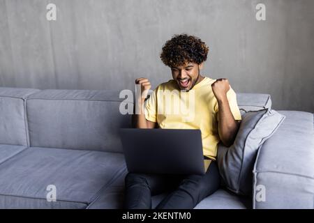 Successful young male lying on the couch while raising hands and using a laptop in the living room Stock Photo